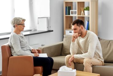 psychology, mental therapy and problem concept - senior woman psychologist taking notes to notebook and sad young man patient at psychotherapy session